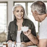 Smiling mature woman talking with man while having coffee. Male and female are spending leisure time during COVID-19 lockdown. They are at home.