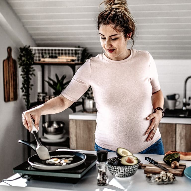 Young Pregnant Woman Preparing Healthy Breakfast in the Kitchen. Cooking eggs and making avocado spread.