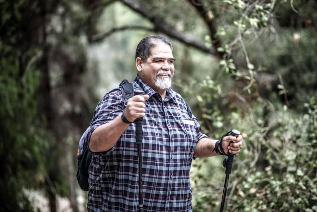A man wearing a plaid shirt hiking in a dense forest with hiking sticks.